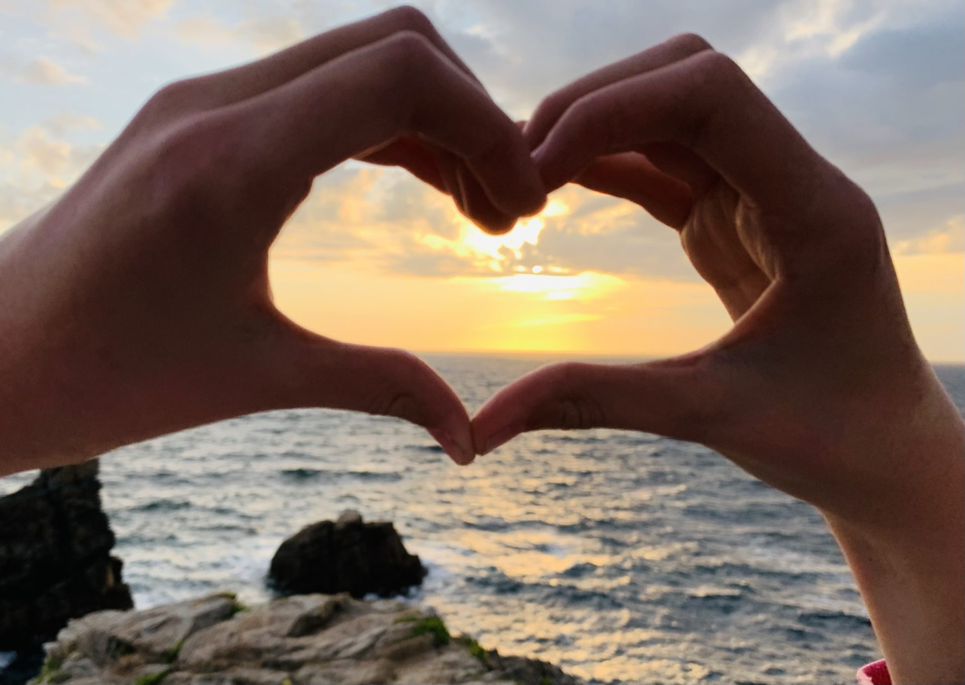 Hände formen ein Herz vor der untergehenden Sonne über dem Meer.